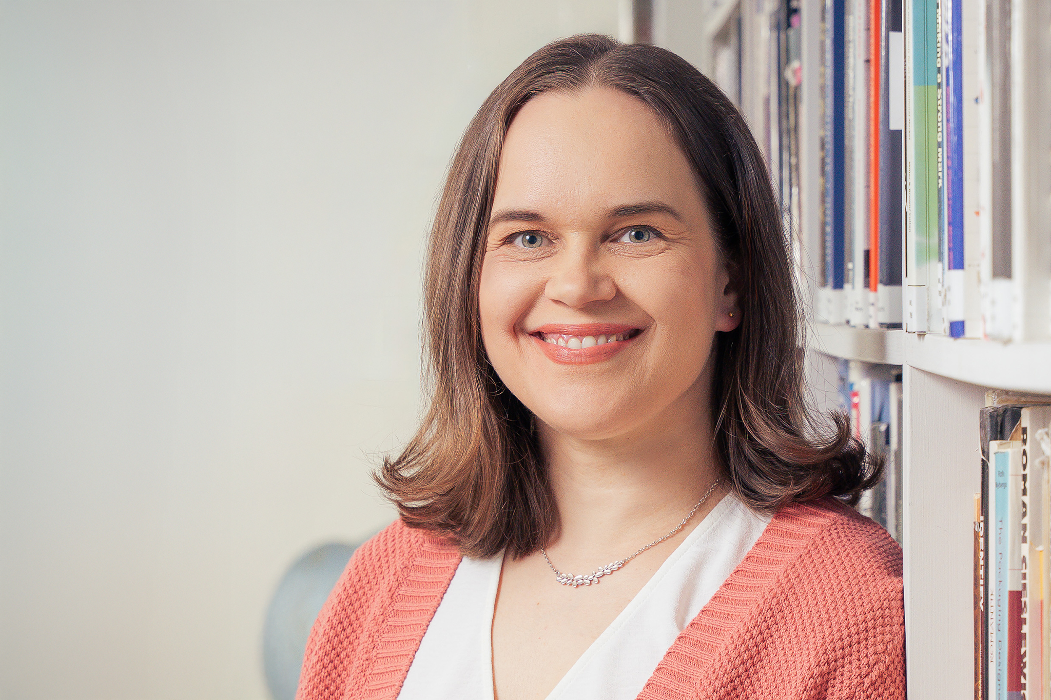 Ruskea hiuksinen henkilö hymyilee. Kirjahylly näkyy taustalla.